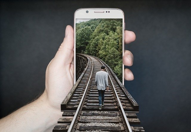 un uomo cammina sui binari di un treno mentre unamano sorregge lo smartphone che mostra l'immagine