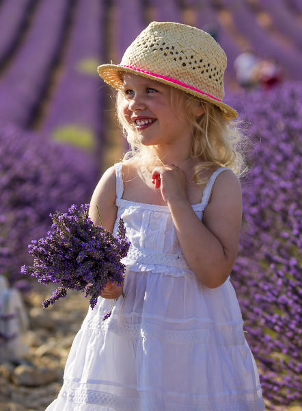 Una bambina bionda con il cappellino in testa sorride con in mano fiori di lavanda in un campo di lavanda